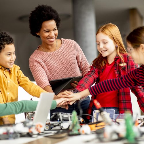 Group of happy kids with their African American female science teacher with laptop programming electric toys and robots at robotics classroom